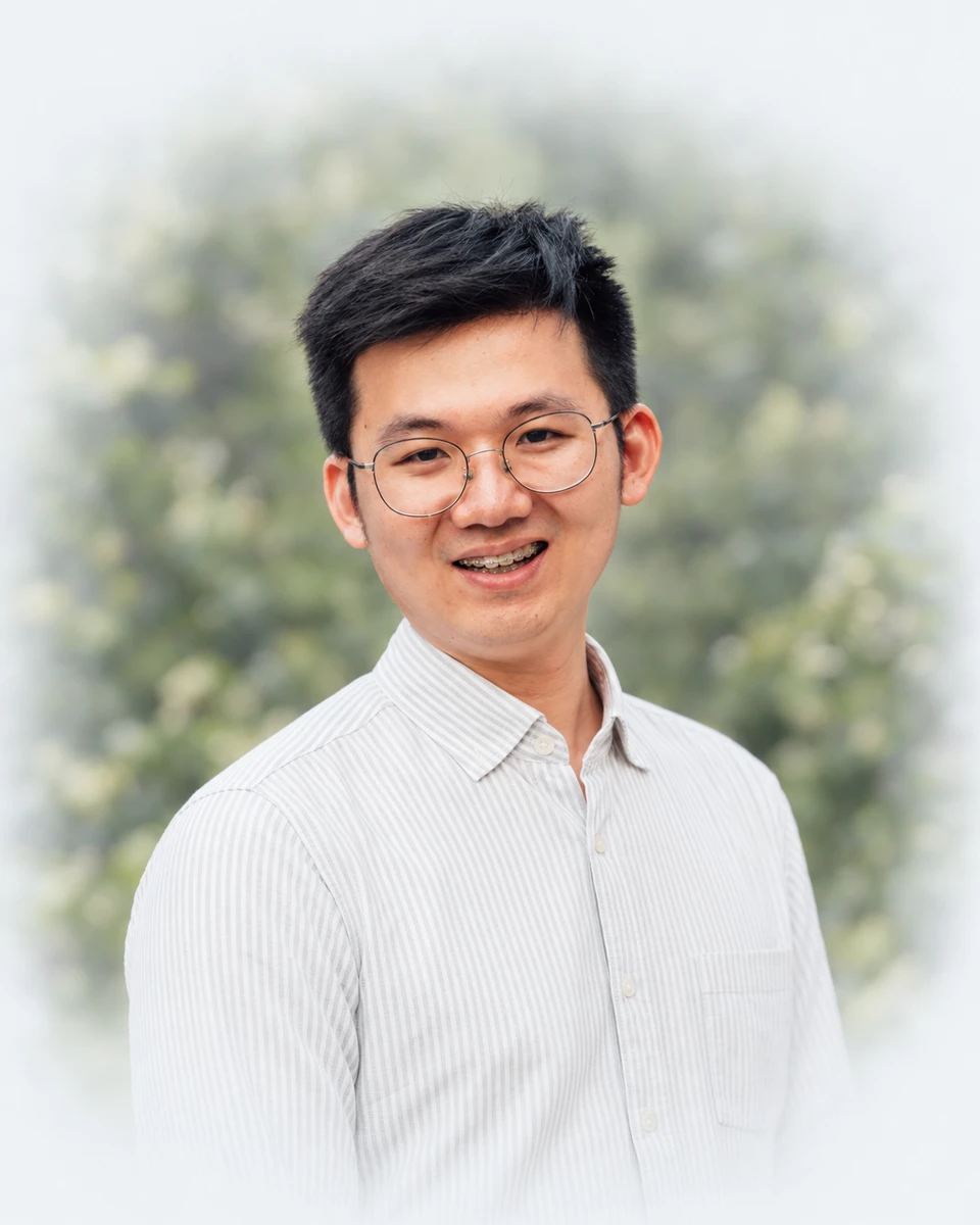 Chong Xian Goh smiling in a pale striped shirt, against softly blurred green foliage.